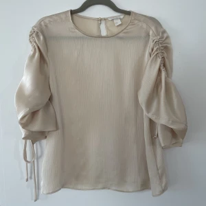 Skimmrig blus från H&M strl 38 - En fin glansig beige blus från hm med snörning på båda ärmar. Går att justera längden på ärmarna. I gott skick 