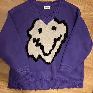 Lila stickad tröja med smileytryck - Har knappt använt, den är rätt så oversize vilket gör det snyggt att ha ett linne eller så under med tanke på att den hänger ner vid axeln. Storlekslappen är bortklippt men den är som sagt oversize