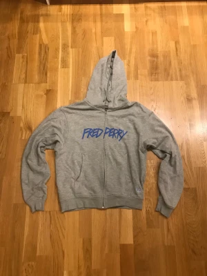 Grå Fred perry zip up hoodie - Säljer min gråa Fred perry zip up hoodie då den är lite för liten för mig, den är i perfekt skick och har ett coolt tryck i graffiti stil. Skriv för fler bilder:kr