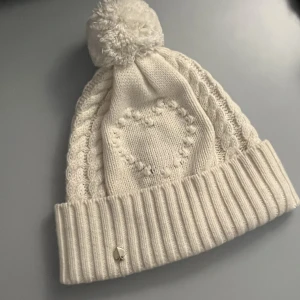 Kate Spade Mössa ”Heart Bobble Beanie” - Modell: Heart Bobble Beanie Färg: Frech Cream (mörk vit) Slutsåld överallt och köptes November 2022.  Provat använda men har alltid hög hästsvans som gör att mössan åker av. Normal storlek.  Utan min hästsvans funkar det: M = 56.5 Inget att anmärka 🩷