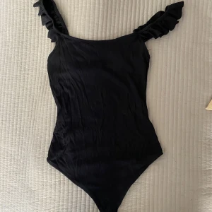 Body/linne med volanger - Gullig body/line med volanger på axelbandet. Sitter tight. Banden är långa men går att justera med säkerhetsnålar. Är i bra skick! 