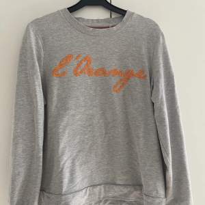 En fin ljusgrå sweatshirt med orangea stenar broderat på framsidan. Storlek M, fint skick.