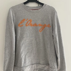 Sweatshirt med stenar - En fin ljusgrå sweatshirt med orangea stenar broderat på framsidan. Storlek M, fint skick.