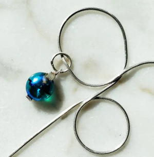 Hängsmycke i djupblått glas - Hängsmycke i metalliskt djupblått glas med handgjord infattning av finsilver (955). Kedjan (Sterlingsilver 925) kan köpas separat. Glaspärlan mäter ca 1 cm.