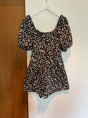 Blommig byxdress - Blommig byxdress med puffärmar. Blommigt svartvitt mönster. Knappar framtill. 