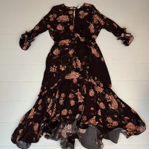 Maxiklänning design Johanna Ortiz - Superfin klänning i fin brun färg med svagt persikofärgade blommor. Lång och lätt utsvängd med volang nedtill och på ärmarna. Använd endast två gånger. Som ny. 