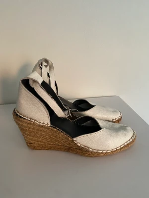 Sandaletter med kilklack - Vita sandaletter med kilklack i skinn och ankelrem i strlk 39. Bra skick, något formade fram men syns inte när man har de på sig.