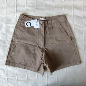 Oanvända khakifärgade shorts från Cubus - storlek 36 - Oanvända khakifärgade shorts från Cubus (modellen heter Zahara) Så snygga och användbara. Högmidjade med stretch, gjorda av ekologisk bomull (98% bomull och 2% elastan). Storlek 36.