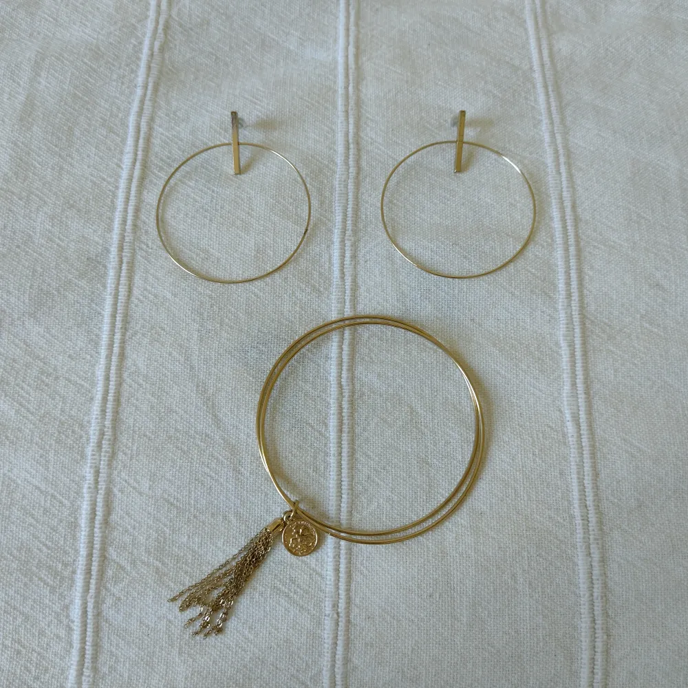 Guldfärgade örhängen i metall. Ringarna är nästan 5 cm i diameter. Något skavda. Armband i form av två tunna ringar och tofs + berlock. . Accessoarer.