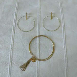 Guldfärgade örhängen i metall. Ringarna är nästan 5 cm i diameter. Något skavda. Armband i form av två tunna ringar och tofs + berlock. 
