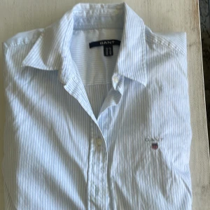GANT dam skjoeta - Ljus blå/vit randig skjorta från GANT i mycket bra skick. Endast använd ett fåtal gånger. Hel. Inga fläckar. Har på vänster arm GANT text i ljust blått. 