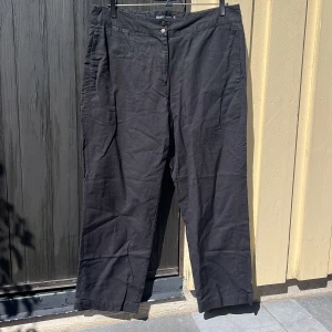 Svarta linnebyxor - Ett par sköna, tunna och svarta byxor. Försedda med en liten framficka, tvåknappsstängning samt dragkedja plus ett litet tygbälte där bak för att justera midjan. 50%linne 50%bomull.