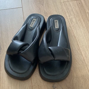 Tofflor/Sandaler  - Svarta tofflor/sandaler med snodd rem framtill som går över foten. Superfina och jättebekväma! Skorna är i fejk-läder.  Inte använda alls mycket så fint skick, dock en liten slitning vid hälen men syns inte när man har på dom! Skriv vid intresse!🖤