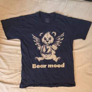 Marinblå t-shirt med en nallebjörn på med ängel vingar. Bra skick förutom ett litet hål vid ryggen som knappt är synligt. Material: 100% bomull