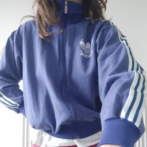 Lilablå vintage adidas jacka - I ett härligt utvättat skick, en lilablå färg med vita detaljer och en ficka på vänster sida. Kort modell i midjan, lappen säger storlek 174 (Frankrike), och i armarna lite kortare men passar mig ändå som är 176cm och strl M