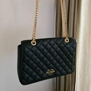 Love moschino axelremsväska - En quiltad axelremsväska som går att ha crossbody eller dubbla remmar över axeln. Kedjan är rörlig. Fint skick. Inga defekter. Rött foder med en liten ficka inuti. En förvaringspåse i tyg följer med. Köpt på nelly.com för 2 år sedan. Fuskläder. Kan skickas spårbart med posten för 66 kr. 