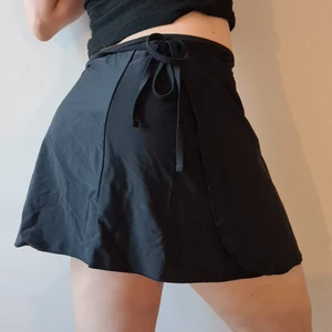 Omlottkjol i baddräktsmaterial - Svart omlottkjol att ha på stranden eller på badhuset. Kjolen är i bikini/baddräktsmaterial (polyamid och elestan) har snörning och kan anpassas i storlek (står dock S på etiketten).