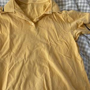 Gul polo T-shirt med två svarta ränder på ärmarna och markerade sömmar, ett broderat M på bröstet. 20kr+frakt