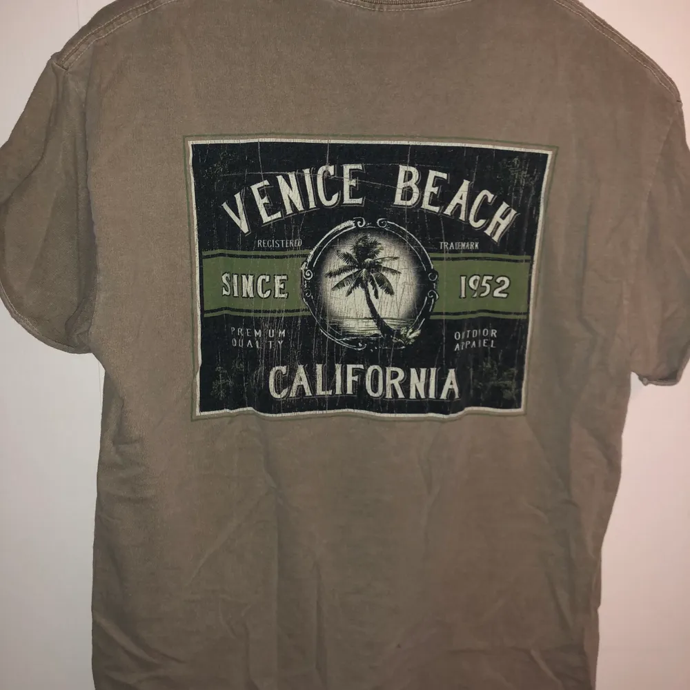 Beige/grå T-shirt med Venice Beach tryck. Litet tryck på höger framsida och stort tryck på ryggen. Uppvikta ärmar. T-shirts.
