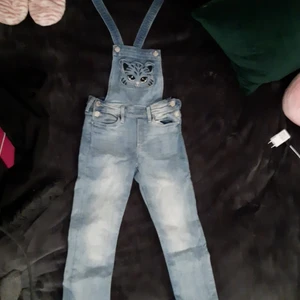 Hängsle byxor för små barn med en katt. - Doma byxorna är inte använda och är i bra skick det är en katt lite längre upp på magen också. Dom köptes för ca 200kr,dom är köpta på H&M och storleken är 128 för 7-8 åringar. Dom är lite blekta vid knäna och mörkare uppåt. Katten är en ficka.