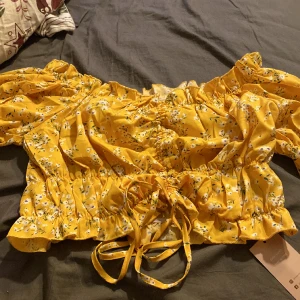 Blommig Croptop Dragsko - En fin, gul och blommig Crop top med en ”dragsko” fram, så man kan förkorta toppen, eller kanske förlänga.