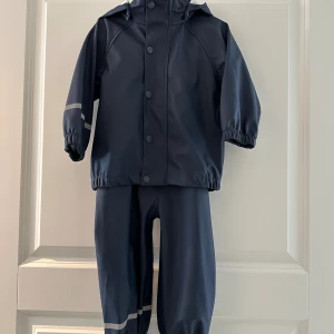 Name It galonkläder regnställ - 80 - Regnställ från Name It storlek 80. Marinblått galon set med byxor och jacka .  Byxorna har hällor och hängslen. Luvan i jackan fästs med knappar.   Mycket fint skick, sparsamt använd. En liten fläck på ärmen som var på den vid köp. 