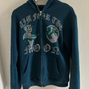 Aim for the Moon Zip Up - Säljer aim for the moon zip up i en grön-blå nyans. Köpt i början av sommar 2023, men har enbart används ett fåtal gånger. Tröjan har inga defekter och alla rhinestones är i bra skick. Vid övriga frågor är det enbart att kontakta oss. 