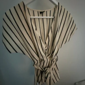 Beige blus - Säljer denna beiga blusen med svarta ränder och ett knytte fram till, kan även knytas bak. Blusen är från ginatricot och är i storlek xs