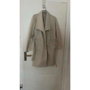 HELT NYTT! Beige tunn kappa från Boohoo - storlek 40 - Säljs den söta fina kappa i beige färg för den är inte min smak. Den är 96 cm från axeln till nedåt. Köpte den ifjol från Boohoo. Hunnit ej använda. Provade endast en gång. Storlek 40