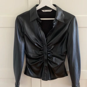 Åtsittande skjorta i skinnimitation  - En svart åtsittande skjorta i skinnimitation o storlek xs. Skjortan är som ny utan prislapp. Så snygg men den har tyvärr inte kommit till användning, skjortan är true to size🥰