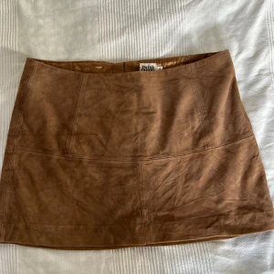 Kort minikjol i mocka - Kort kjol till sommarens fester och tillfällen. Material i mocka. Storlek 36, passar dig som har en omkrets på ca 84 cm. Sliten look som förhöjer hela outfiten. Längd: 35 cm. Dragkedja på baksidan av kjolen.  Märke: TWIST AND TANGO