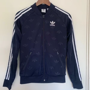 Blå Printed Adidas Zip-Up - Blå Adidas zip-up med printad logga. Storlek XS herr. Fint använt skick. Två väldigt små fläckar på en av de vita ränderna på armarna, utöver det inga anmärkningar.