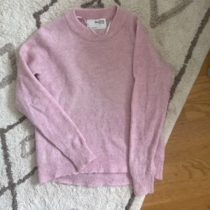 Rosa stickad tröja från Selected Femme - En mjuk och bekväm rosa stickad tröja från Selected Femme. Tröjan har långa ärmar och ribbstickade muddar vid ärmslut och nederkant. Perfekt för kyligare dagar.