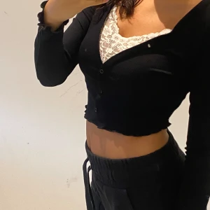 Svart croppad topp med spetsdetaljer - En svart croppad topp med långa ärmar och spetsdetaljer vid halsringningen. Toppen har knappar framtill och en åtsittande passform.