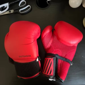 Röda boxningshandskar från Outshock - Ett par röda boxningshandskar från Outshock med svart kardborreband för justering. Handskarna har en vadderad yta och meshmaterial på insidan för ventilation.