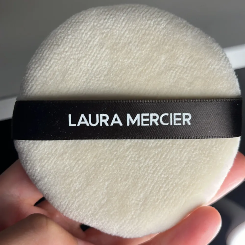 En rund, vit puderpuff från Laura Mercier. Oanvänt pga fel storlek. Puffen har en mjuk yta och ett svart band med märkesnamnet tryckt i vitt. . Skönhet.