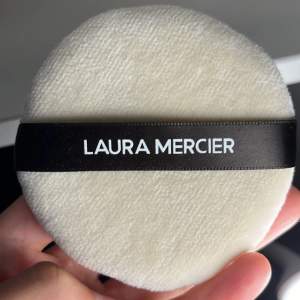 En rund, vit puderpuff från Laura Mercier. Oanvänt pga fel storlek. Puffen har en mjuk yta och ett svart band med märkesnamnet tryckt i vitt. 