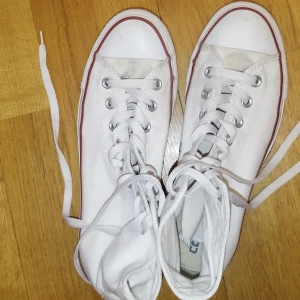 Vita höga sneakers från Converse - Ett par vita sneakers från Converse med snörning och den klassiska Converse All Star-logotypen på sidan. Skorna har en vit sula med en röd rand. Den är sparsam använd. Pris kan sänkas vid snabb affär.