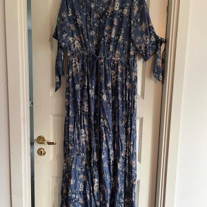 Blommig långklänning - En vacker långklänning med blommönster i olika nyanser av beige och vitt på en blå bakgrund. Klänningen har en knytning i midjan och korta ärmar med knytband. Den är tillverkad i ett lätt och luftigt material, perfekt för sommar och vår. Måste sys lite i midjan men inget hål, se bild
