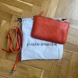 Orange väska - Clutch från Carin Wester. Aldrig använd. Dustbag medföljer. Orange, skinnimitation.