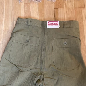 Gröna 90-tals byxor utsvängda  - Vintage utsvängda byxor från Dobber, grönbeige färg. Helt nya med lapp kvar, endast legat på lager.  Obs det är rålängd, de är alltså inte fållade utan bör sys upp i ens egen längd. Märkta size 30, inseam 36.  Mått cm: Midja: 35 Höft: 43 Ben: 88  