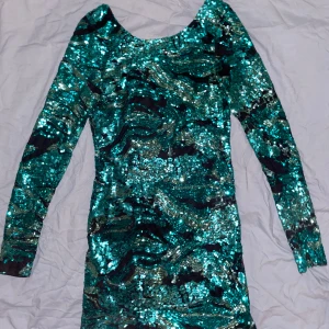 Glittrig svart/grön cocktailklänning från Rebecca Stella for Nelly. - Svart/Grön Klänning med öppen rygg, en festlig cocktail klänning.  Använd några gånger så det kan ha tappat lite glitter på några ställen men det är knappast synligt.  Kan inte ha den längre så sedan dess har den bara legat i garderoben.