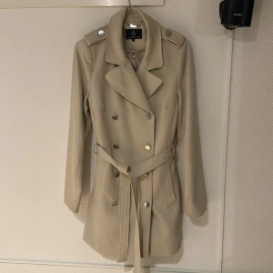 Beige Vårkappa 38, väldigt bra skick - Fräsch kappa i mjukt halvgrovt tyg. Kappa , trenchcoat. Använd mindre än 5 gånger. En normal 38 så något stor för mig som ofta har 36 fast kan behöva 38 över axlarna. Knälång Fina blanka silverknappar. Märke STOCKH LM