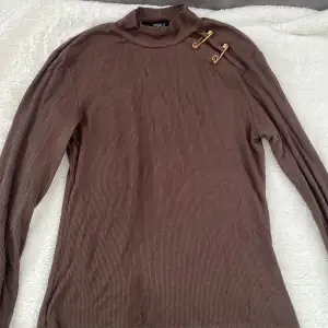 Fin ribbad brun tröja med kort hals och fin detalj vid vänster axel med fake säkerhetsnålar med detaljer i guld. Ej använd.