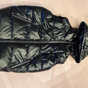 Glansig svart dunväst - Dunväst inyskick använd fåtal gånger. Fickor i sidan, luva. Stl 34/36