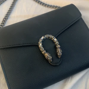 Väska  - Svart axelremsväska med orm på, många fack i väskan även en korthållare, använd mycket, trasig dragkedja på insidan men går att fixa lätt 