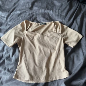 Beige ribbad topp - En beige ribbad topp med korta ärmar och en fyrkantig halsringning. Toppen har en figurnära passform och är tillverkad i ett stretchigt material.