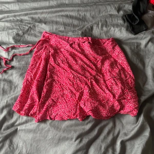 Röd kortkjol med blommigt mönster - En röd kortkjol med ett blommigt mönster. Kjolen har en omlott-design och knytband i midjan.