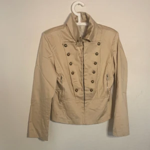 Beige jacka - Jacka med axelvaddar och cool knäppning. Saknar en knapp vid höger sida (framifrån) på kragen men användbar ändå