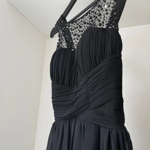 Balklänning svart  - Svart slät balklänning med ett fint fall och ett nät av pärlor över bröstet. Vadderade kupor. Fungerar både till bal och som exempelvis bröllopsgäst. Användes endast en gång. Som ny! Storlek 40, ca 150 cm lång från axelbanden till botten.